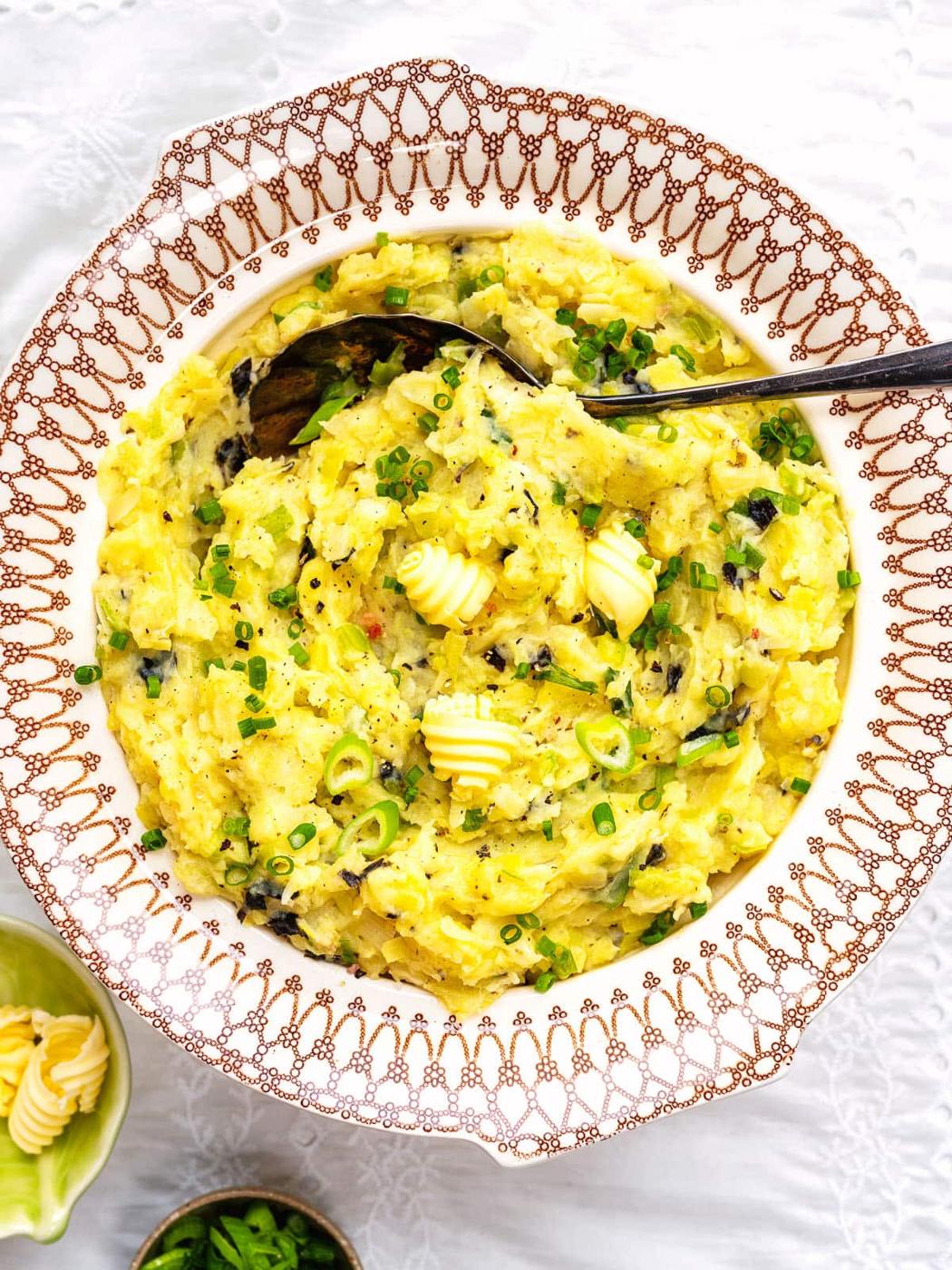 A decorative bowl filled with colcannon—creamy mashed potatoes blended with greens, garnished with chopped chives, black pepper, and butter curls. A serving spoon rests inside, with a small dish of extra butter beside the bowl.