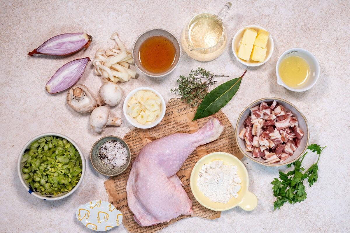 Raw ingredients for a recipe are arranged on a countertop, including a chicken leg, chopped celery, garlic, mushrooms, butter, flour, shallots, bay leaf, thyme, parsley, salt, pepper, diced bacon, oil, wine, and broth.