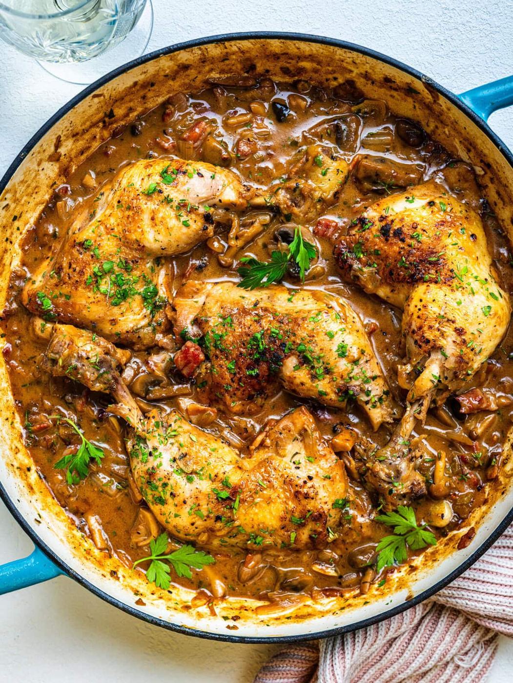 A large skillet filled with golden-brown chicken legs simmering in a creamy, herbed mushroom and tomato sauce, garnished with chopped parsley. A glass of water and a striped towel are nearby.