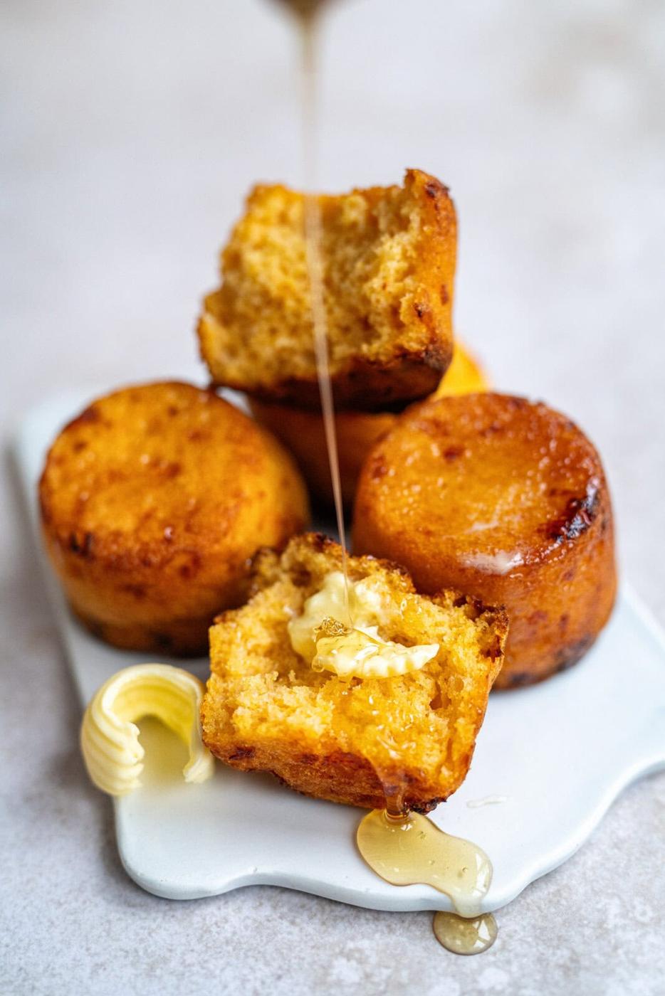cornbread muffins on a white ceramic board, one muffin is split open with butter and drizzled with honey