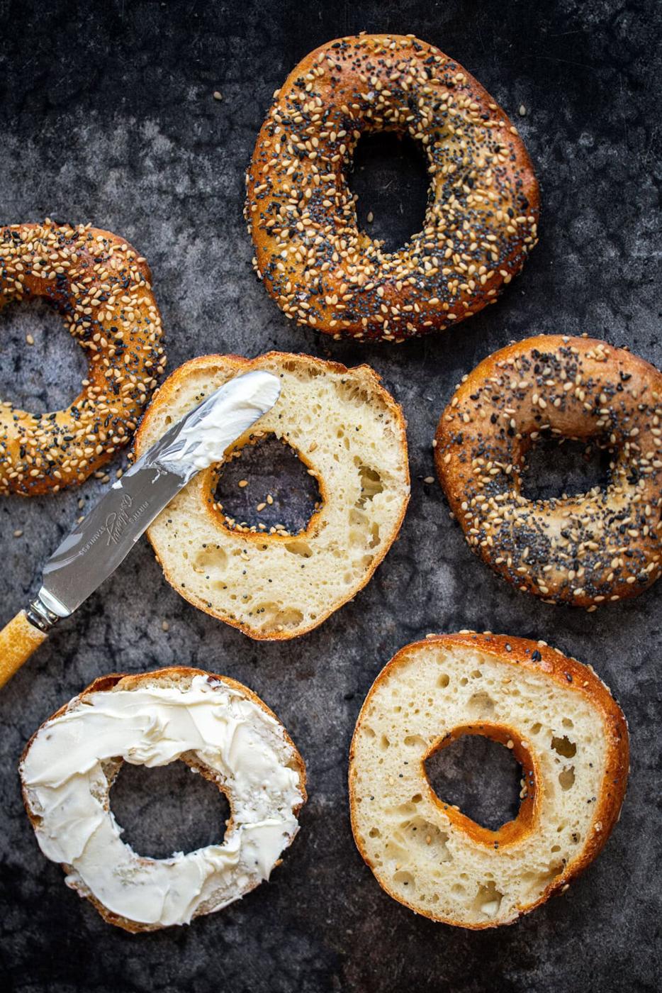 A group of cottage cheese bagels, some with sesame and poppy seeds, are arranged on a dark surface. One sliced bagel is spread with cream cheese, with a knife resting beside it.