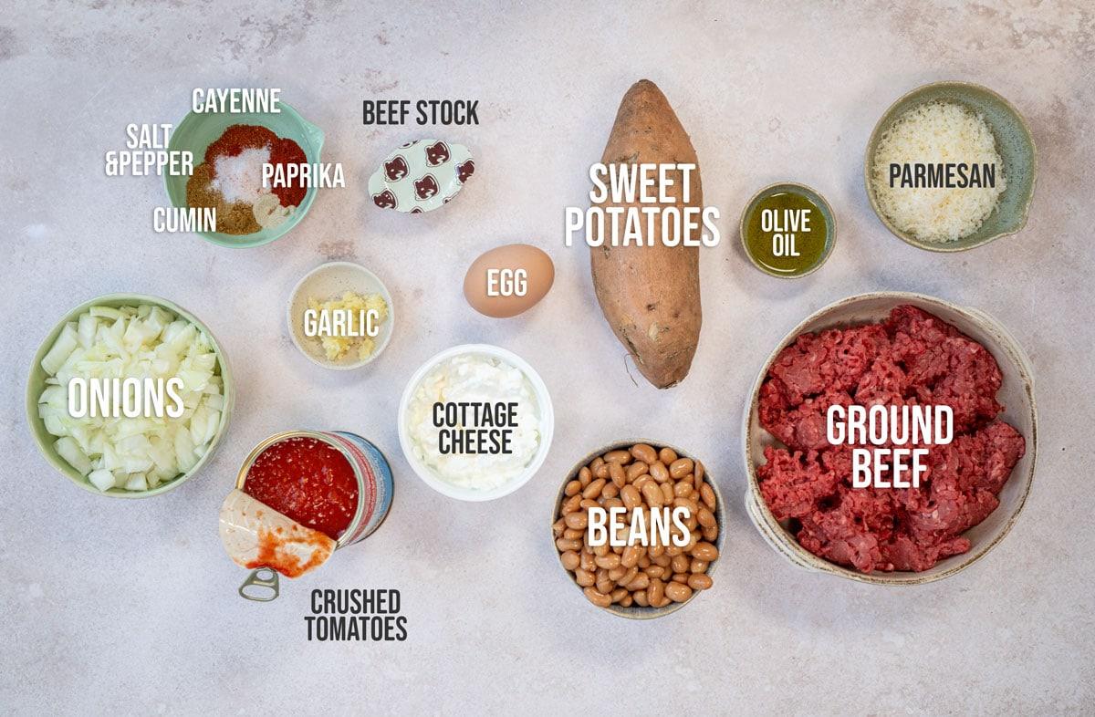 Ingredients arranged on a surface include ground beef, sweet potatoes, beans, cottage cheese, onions, crushed tomatoes, egg, garlic, parmesan, olive oil, beef stock, and spices such as cayenne, paprika, cumin, salt, and pepper.