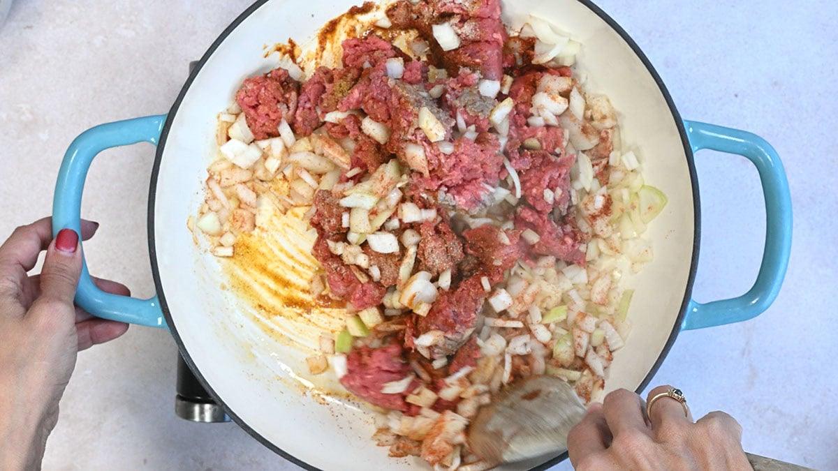 Top view of a person cooking ground beef and diced onions in a large blue-handled saucepan. The person is stirring the mixture with a wooden spatula, and raw pink meat is seen among the browned edges and softened onions.