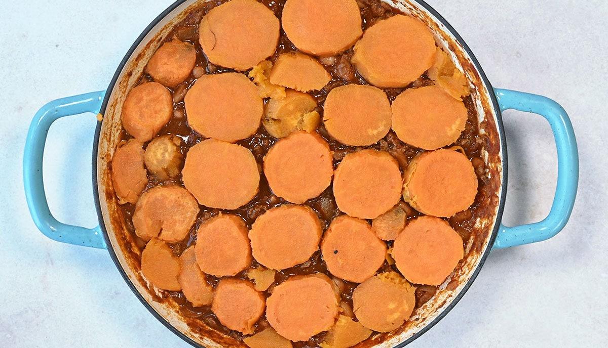 A round blue dish filled with a baked casserole. The top layer features neatly arranged, circular slices of sweet potatoes in a golden-brown sauce, giving the dish a warm and appetizing appearance.