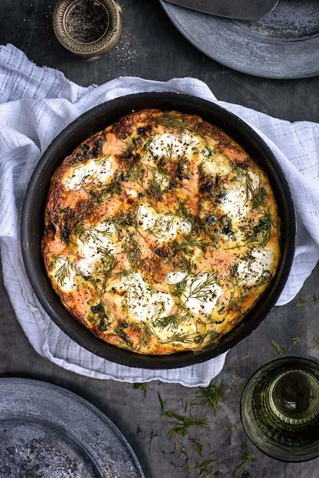 A freshly baked frittata in a round black dish on a white cloth. The frittata is topped with melted cheese, herbs, and crispy edges. Scattered dill sprigs are visible, with a blurred plate, fork, and pepper on the side.