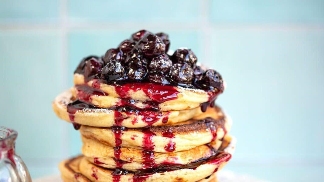 A stack of cottage cheese pancakes topped with glossy, dark berries and drizzled with berry syrup sits on a white marble board. The background features light blue tiles, and theres a glimpse of a jar filled with berries beside the pancakes.