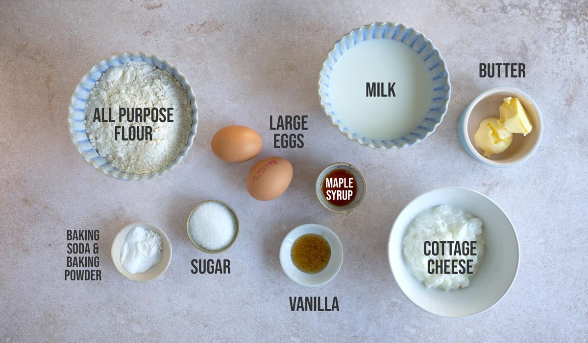 Ingredients for pancakes are arranged on a surface: all-purpose flour, baking soda and powder, sugar, large eggs, vanilla, maple syrup, milk, butter, and cottage cheese. Each item is labeled with text.