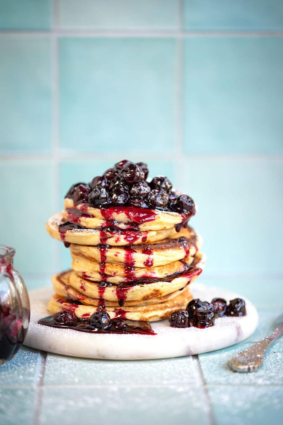 A stack of cottage cheese pancakes topped with glossy, dark berries and drizzled with berry syrup sits on a white marble board. The background features light blue tiles, and theres a glimpse of a jar filled with berries beside the pancakes.