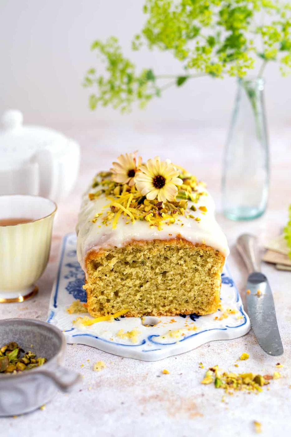 A Lemon Zucchini Bread With Cream Cheese Glaze, garnished with pistachios and edible flowers, sits on a decorative plate. Nearby are a cup of tea, a knife, and a jar with green flowers on the light, airy table.