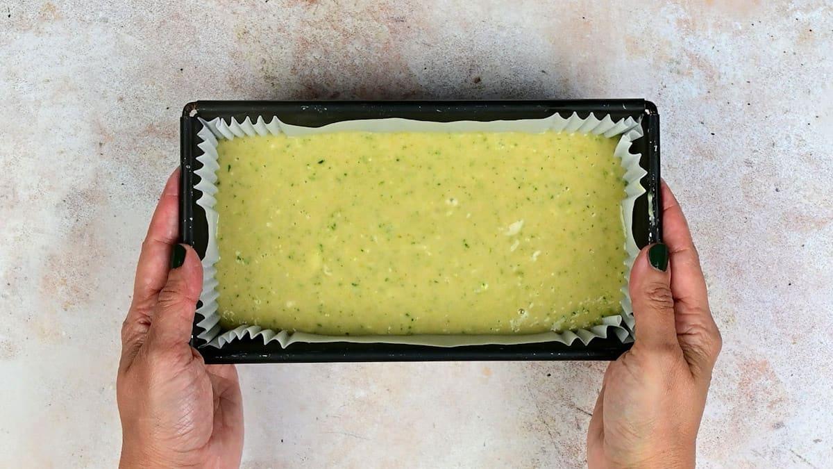 A person holds a rectangular baking tin lined with parchment paper, filled with uncooked Lemon Zucchini Cake With Cream Cheese Glaze batter, speckled with green, likely zucchini, over a light textured surface.