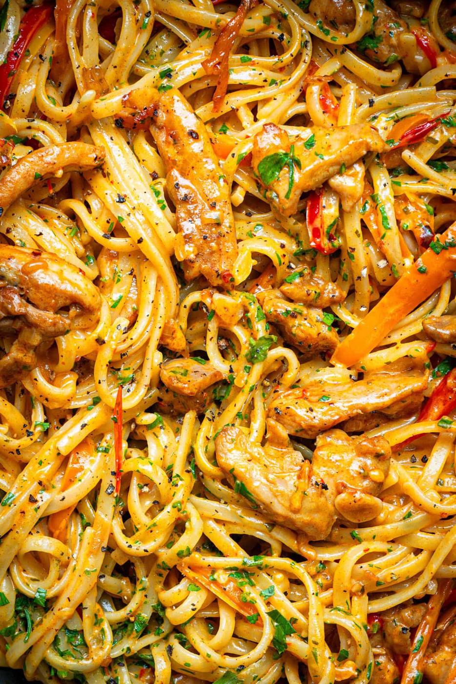 A close-up of creamy pasta with strips of chicken, sliced red and yellow bell peppers, and chopped parsley, all coated in a rich sauce. The dish appears flavorful and freshly prepared.