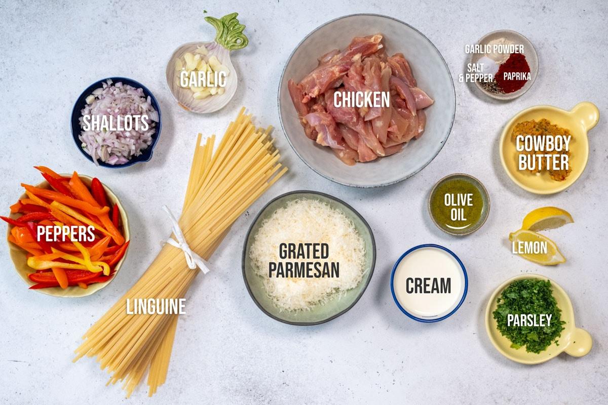 Top-down view of ingredients for a pasta dish: shallots, garlic, chicken, cowboy butter, paprika, salt, pepper, garlic powder, olive oil, lemon wedges, parsley, cream, grated parmesan, linguine, and sliced red peppers.