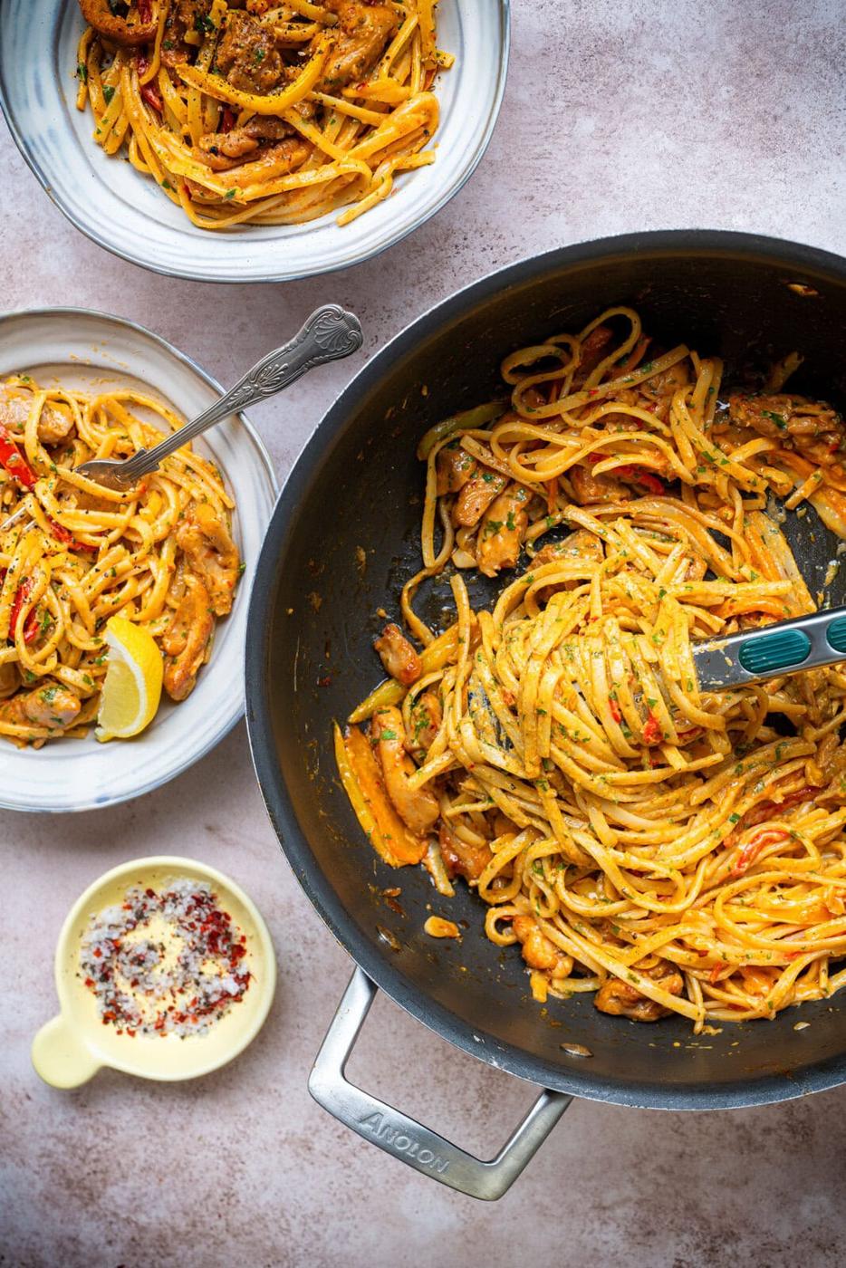 A pan filled with creamy pasta and vegetables sits on a table beside two bowls of served pasta and a small dish of grated cheese with pepper flakes. A fork is twirling pasta in the pan.