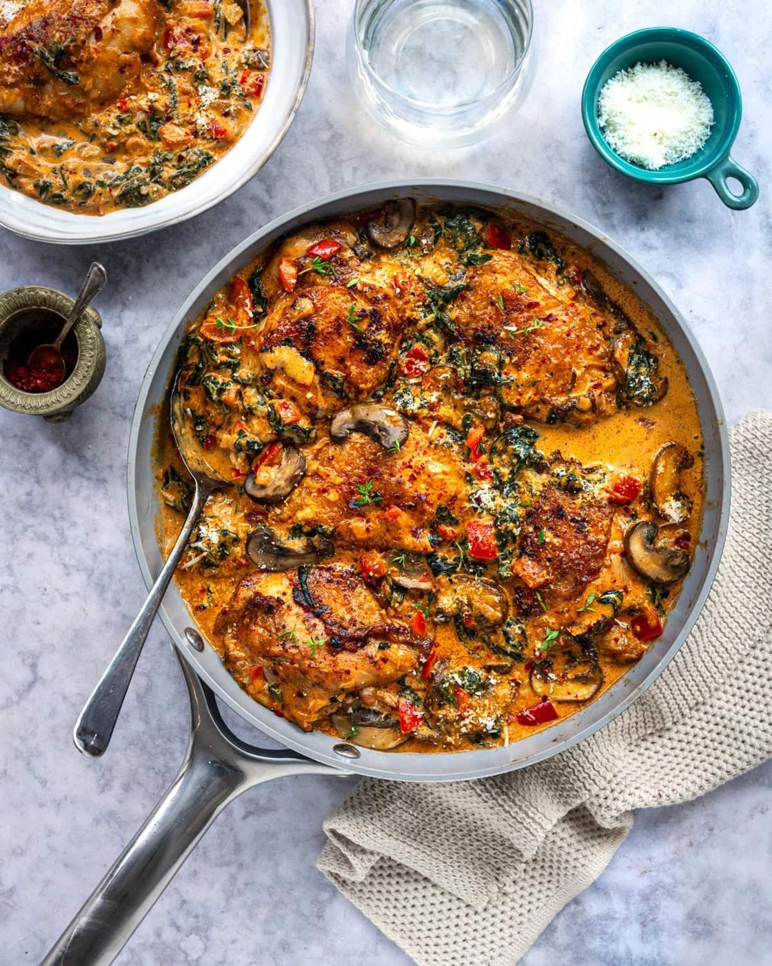 A skillet filled with creamy paprika chicken thighs, mushrooms, spinach, and red peppers in a rich sauce. A spoon rests in the skillet. Nearby are a bowl of grated cheese, a glass of water, and a textured beige cloth.