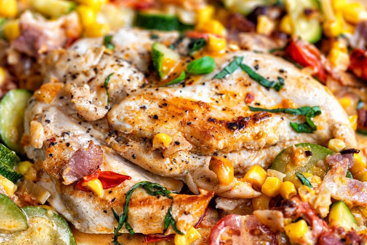 close up on chicken breasts cooked in a creamy sauce with bacon, corn, zucchini and tomatoes