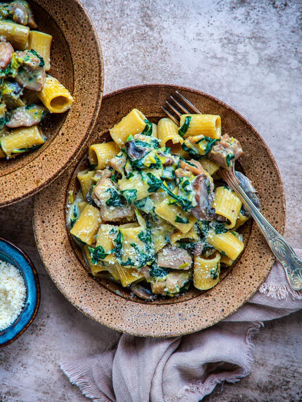 A bowl of rigatoni pasta mixed with mushrooms, spinach, and a creamy sauce, topped with grated cheese. Another bowl of pasta and a small dish of cheese are nearby on a textured surface with a cloth napkin.