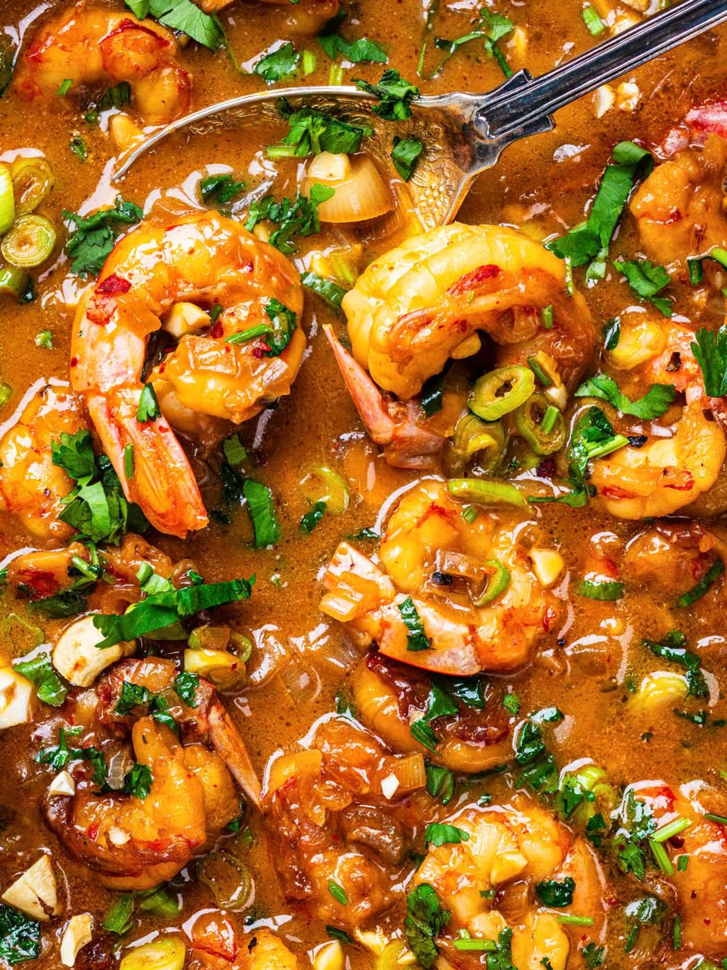 Close-up of a flavorful shrimp dish in a rich, brown sauce, garnished with chopped green onions and fresh herbs, with a spoon partially visible in the background.
