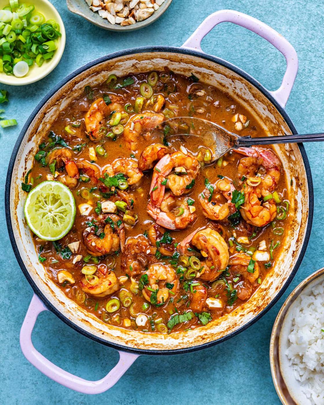 A pot of creamy coconut shrimp garnished with chopped green onions and lime slices, with a serving spoon inside. Surrounding bowls contain rice, chopped nuts, and sliced green onions on a blue background.
