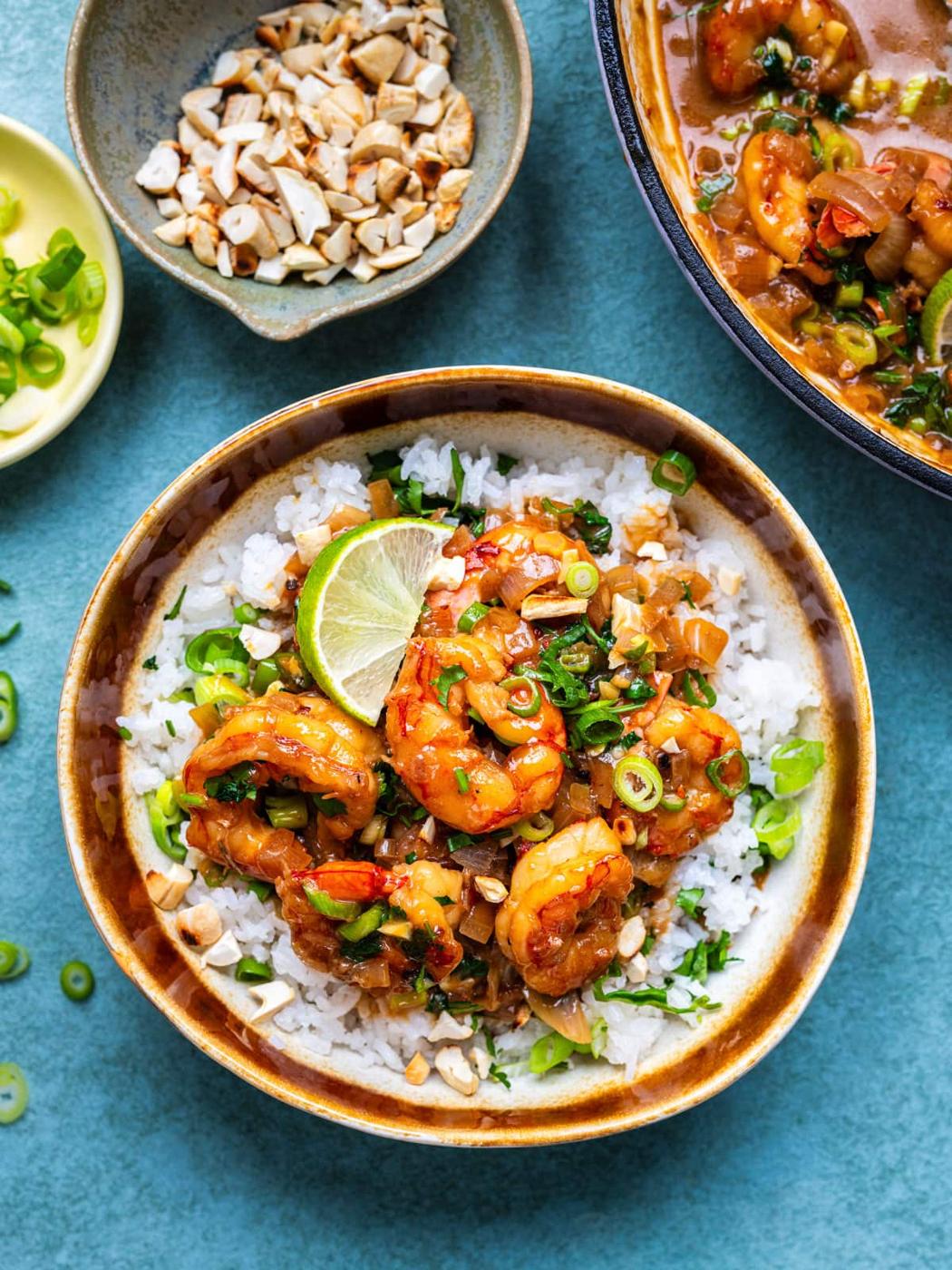 A bowl of white rice topped with saucy coconut shrimp, chopped green onions, cilantro, and a lime wedge. Nearby are bowls of chopped nuts, sliced green onions, and more shrimp in sauce.