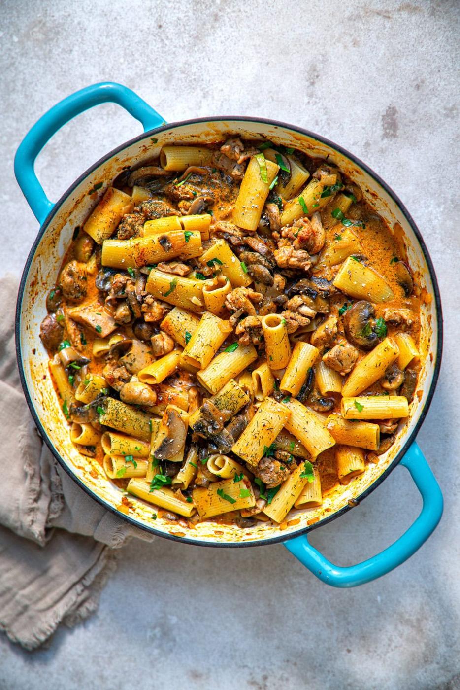A blue-handled pot filled with rigatoni pasta, mushrooms, and chicken in a creamy sour cream sauce. Fresh parsley is sprinkled on top for garnish. A beige cloth is beside the pot on a textured, neutral background.