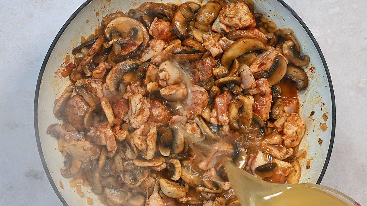 A pan filled with sliced mushrooms and chunks of chicken being cooked. A liquid, likely broth, is being poured into the pan, creating steam. The ingredients appear to be sautéing, creating a savory dish.