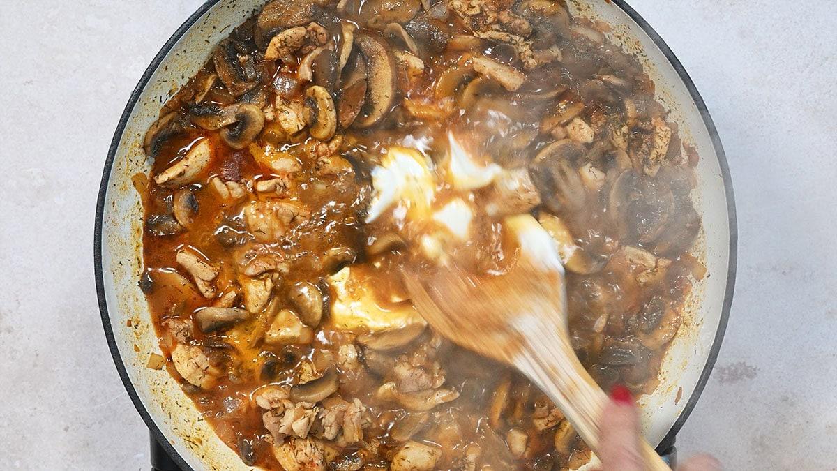 Hand stirring mushrooms, chicken, and a creamy sauce in a skillet with a wooden spoon.