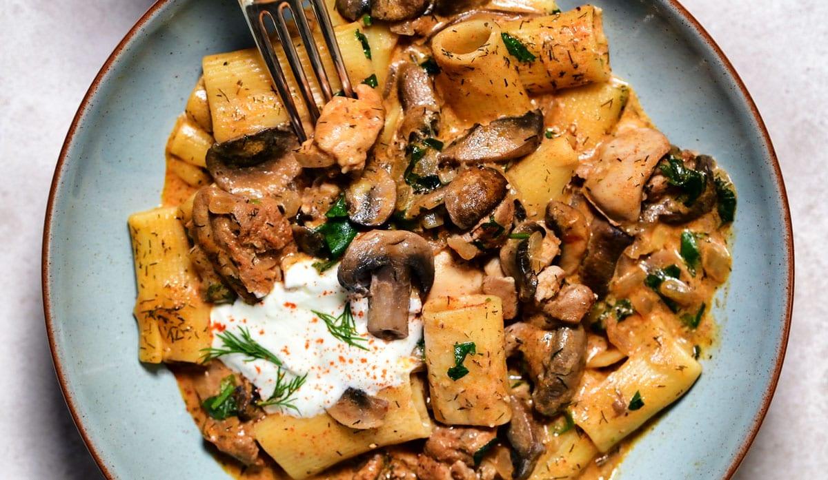 A bowl of pasta with rigatoni, mushrooms, and chicken in a creamy sauce. Garnished with fresh herbs and a dollop of sour cream. A fork rests on the side of the dish.