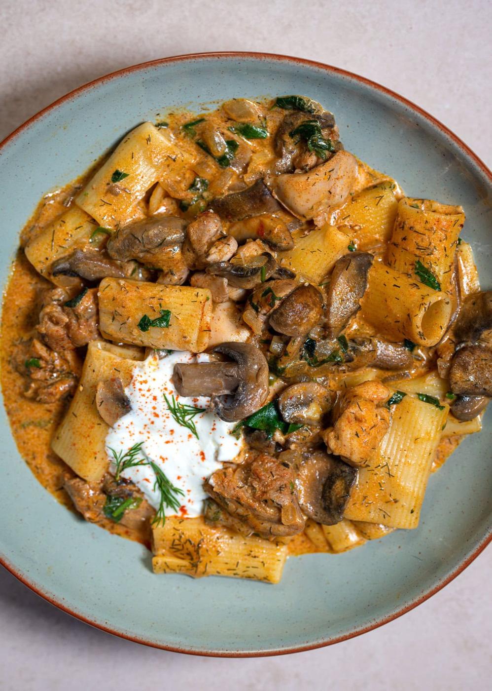 A plate of rigatoni pasta with chunks of chicken, mushrooms, and fresh herbs in a creamy sauce, garnished with a dollop of sour cream and a sprinkle of paprika.