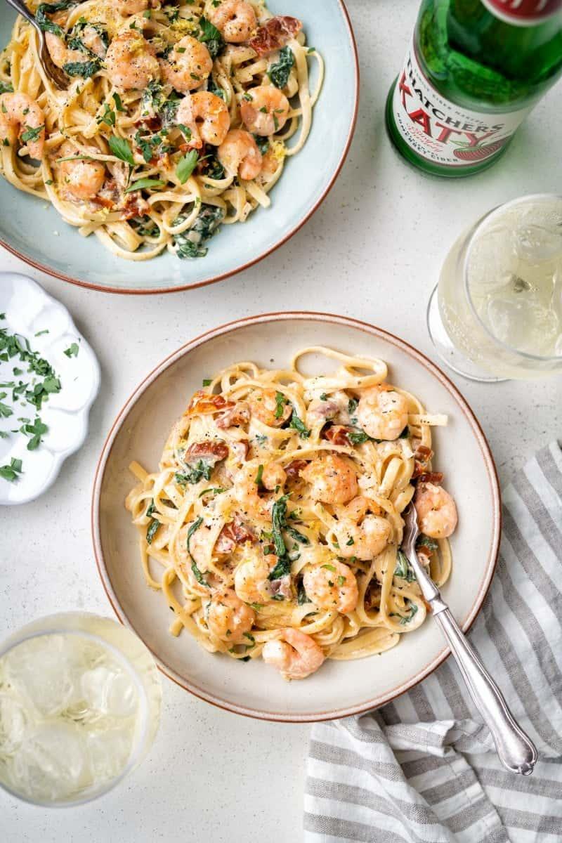 Two bowls of creamy Tuscan shrimp pasta with spinach and sun-dried tomatoes sit on a white table beside a glass of ice, chopped herbs on a small plate, a striped napkin, and a bottle of sparkling water.