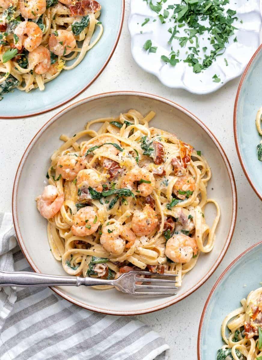 A bowl of creamy Tuscan shrimp pasta with sun-dried tomatoes, spinach, and herbs, garnished with grated cheese, sits on a plate with a fork. Other similar plates and a dish of chopped parsley are nearby.