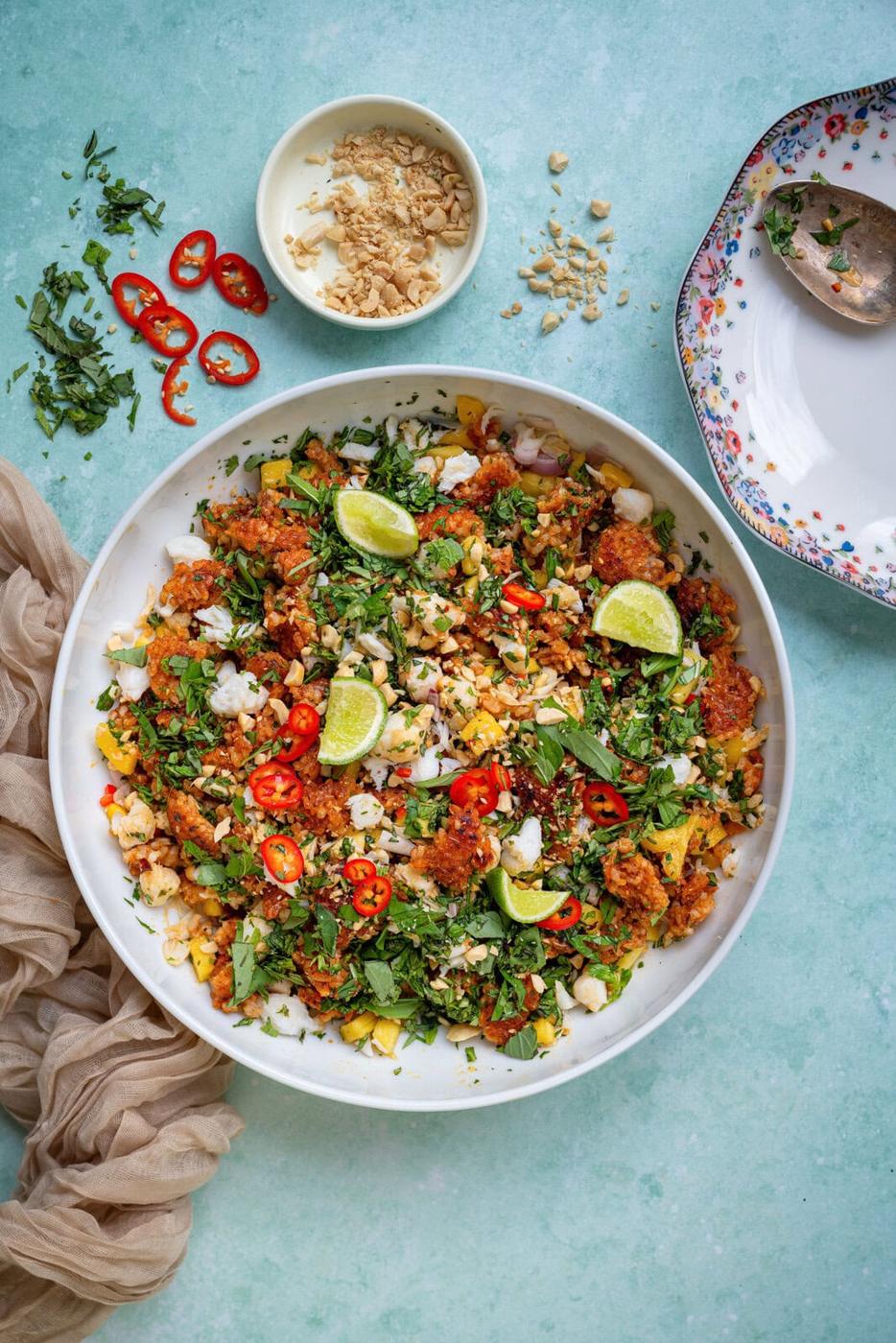 A colorful crispy rice salad in a large white bowl, topped with lime wedges, chopped herbs, peanuts, and sliced red chilies, sits on a light blue surface beside a floral plate, spoon, and small bowl of chopped peanuts.