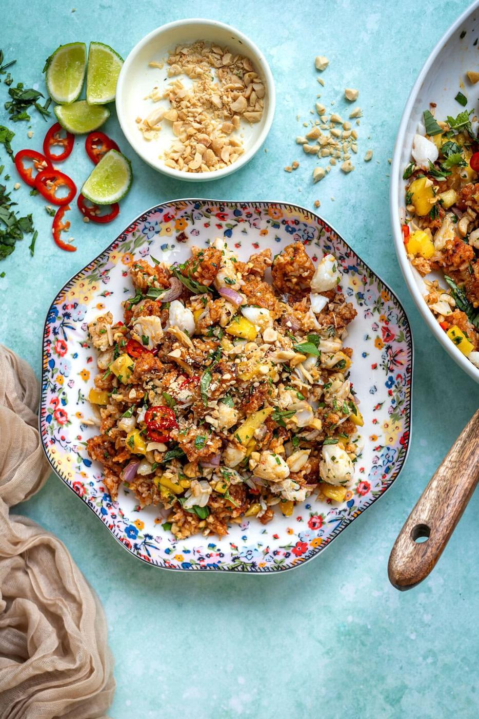 A colorful plate crispy rice with vegetables, tofu, red chili, fresh herbs, and crushed peanuts, surrounded by lime wedges, chopped peanuts, and a cooking pan on a light turquoise background.