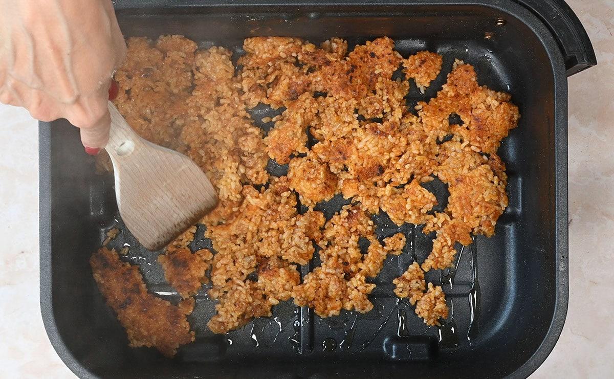 A hand uses a wooden spatula to scrape browned, crispy rice from the bottom of an air fryer basket, revealing small clumps of golden, toasted rice.
