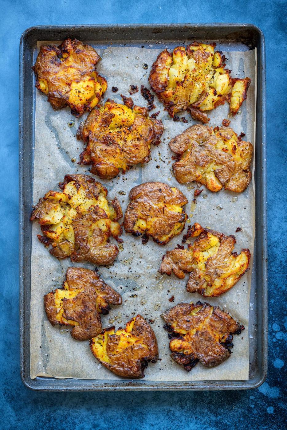 A baking sheet with nine golden-brown smashed potatoes, seasoned with herbs and spices, on parchment paper. The bluish surface beneath the tray adds contrast to the crispy potatoes.