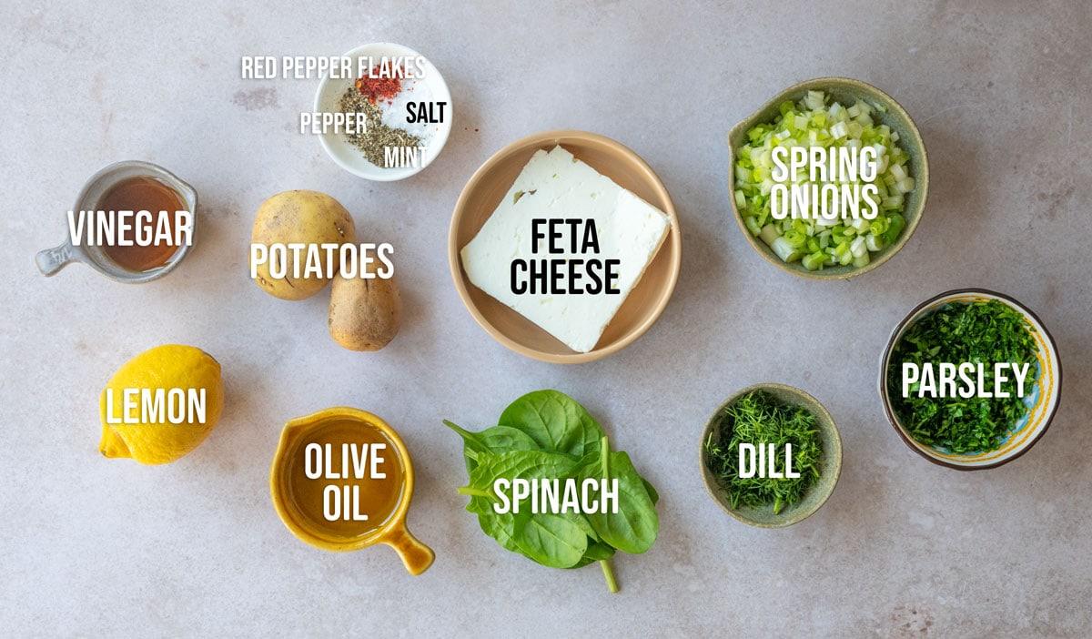 A variety of labeled ingredients on a table includes vinegar, lemon, olive oil, potatoes, spinach, feta cheese, spring onions, parsley, dill, and seasonings such as red pepper flakes, salt, and dried mint.