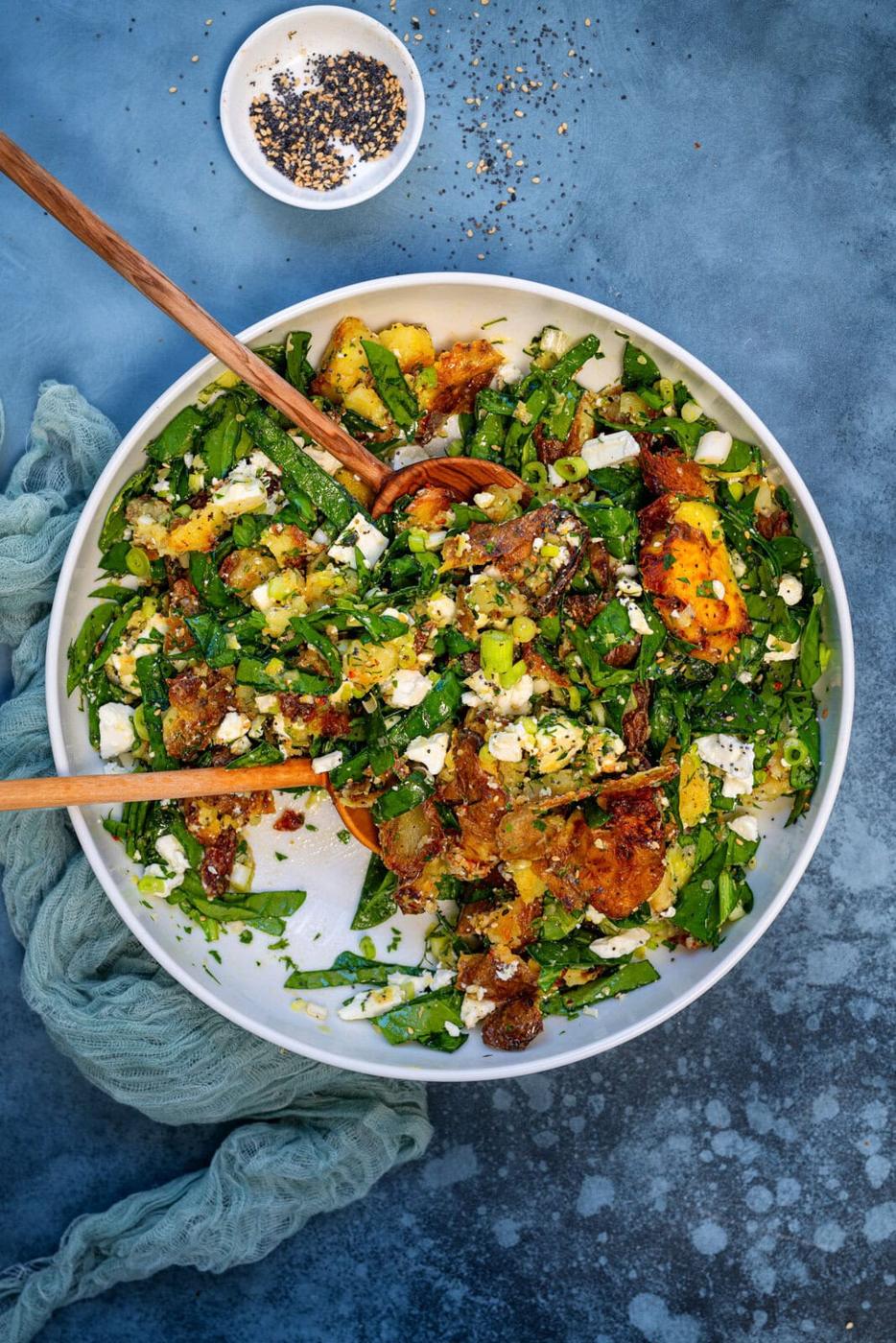 A vibrant salad with spinach, smashed potatoes, feta cheese, served in a white bowl on a blue surface. Wooden tongs lie in the bowl, and a small dish with mixed seeds is nearby. A light green cloth is beside the bowl.