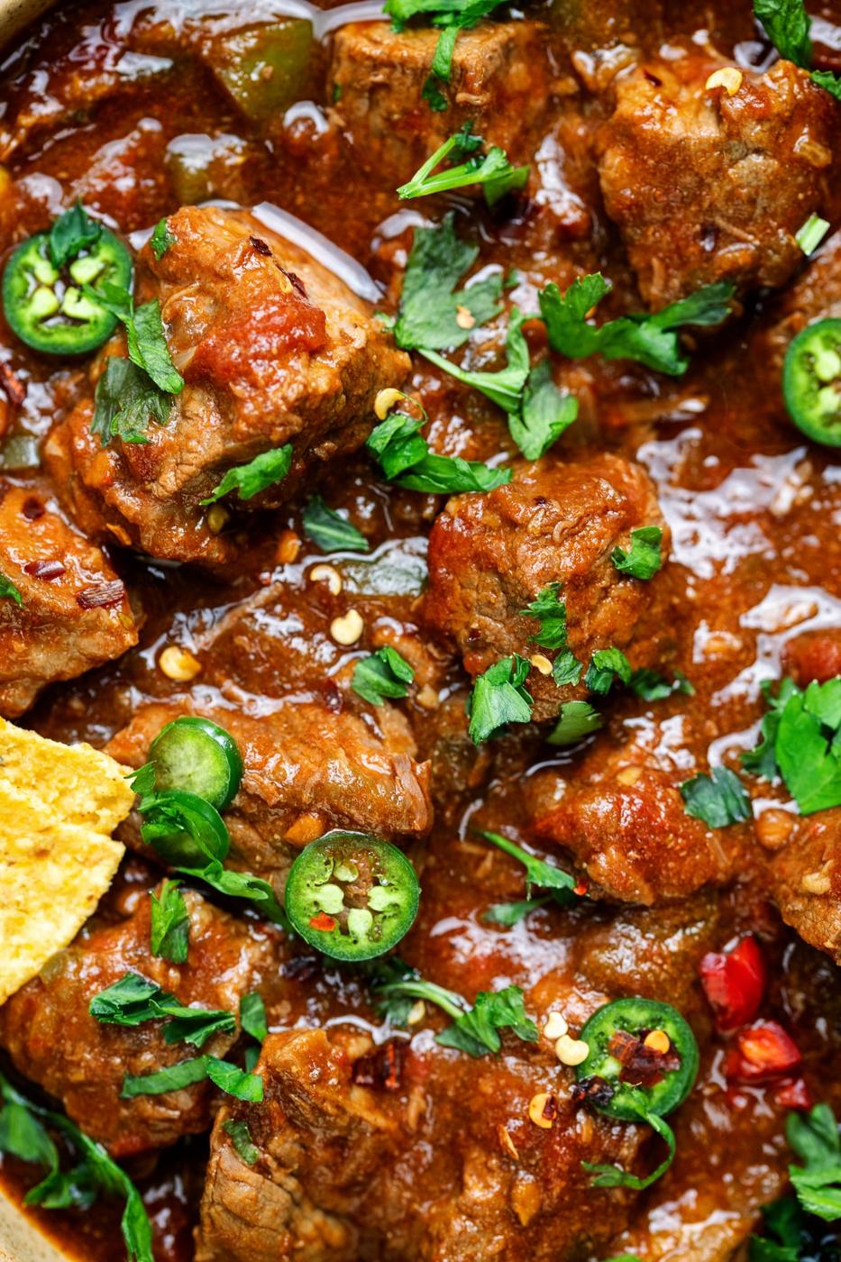 Close-up of spicy Crock Pot carne guisada beef chili with chunks of tender meat in a rich, red sauce, garnished with sliced green jalapeños, fresh cilantro, and a tortilla chip on the side.