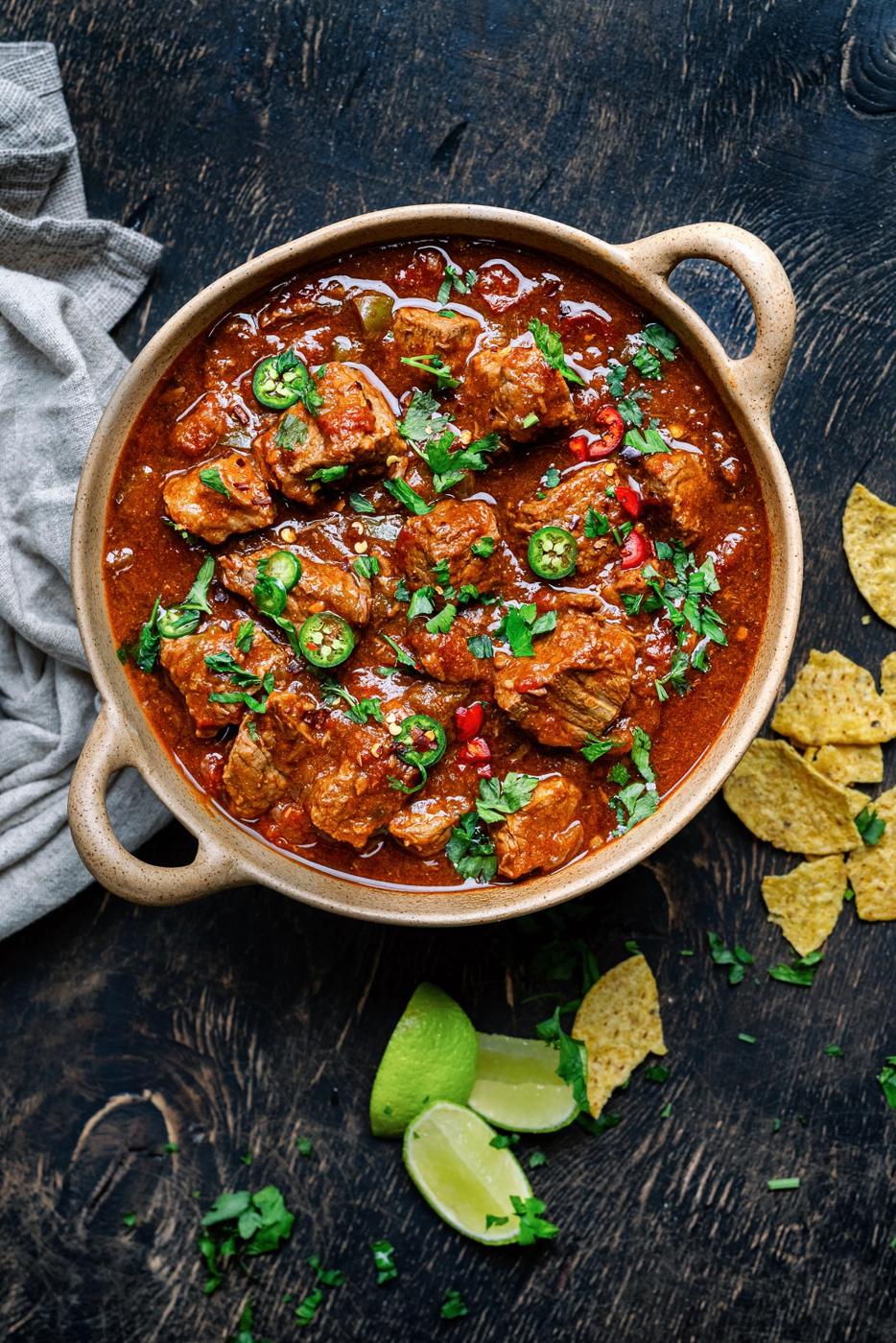 A bowl of spicy Crock Pot carne guisada beef stew garnished with chopped cilantro and sliced green chilies, surrounded by lime wedges, tortilla chips, and a gray napkin on a dark wooden surface.
