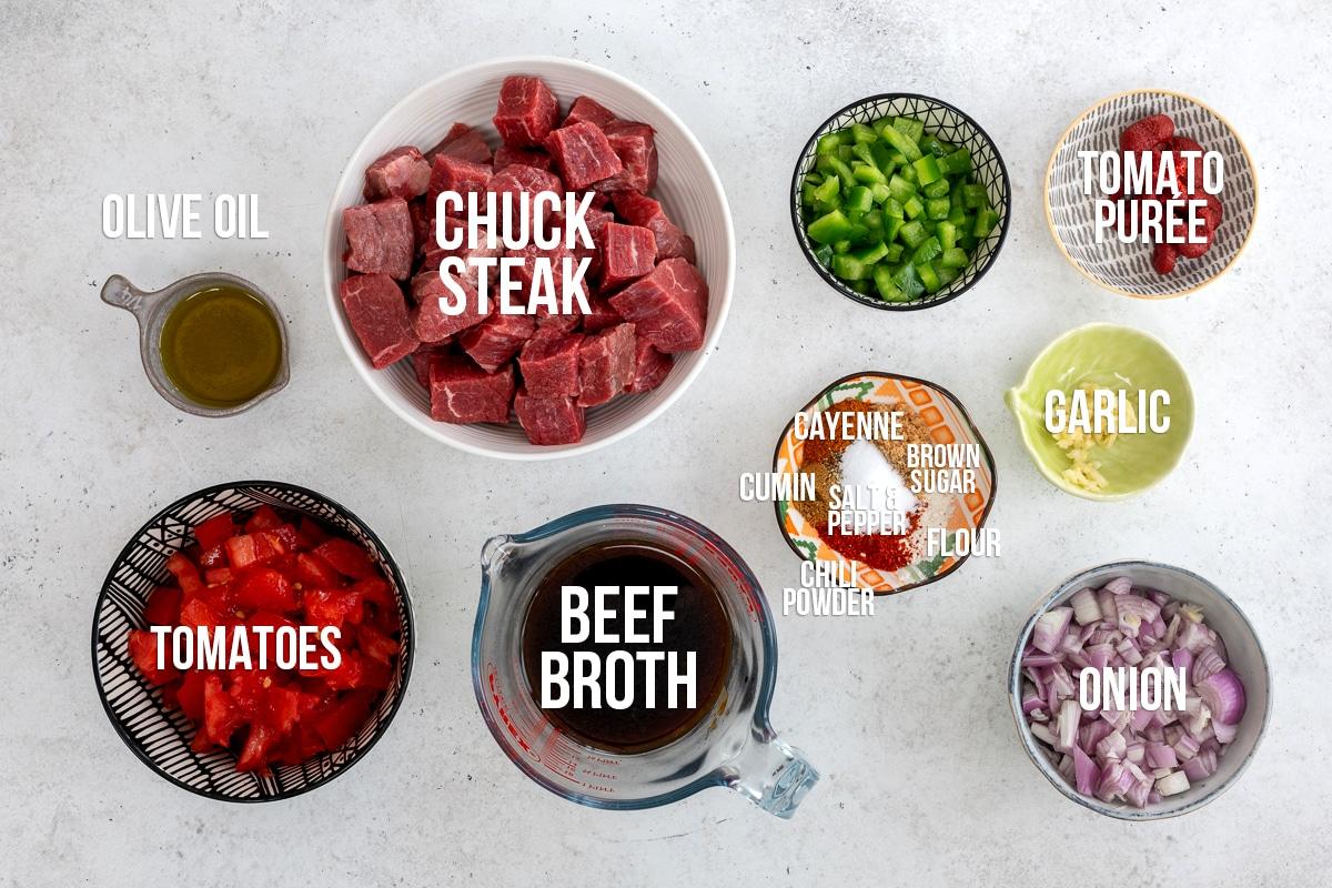 Top-down view of chili ingredients in bowls: cubed chuck steak, chopped tomatoes, beef broth, onion, green bell pepper, tomato puree, garlic, olive oil, and spices—perfect for making Crock Pot carne cuisada.