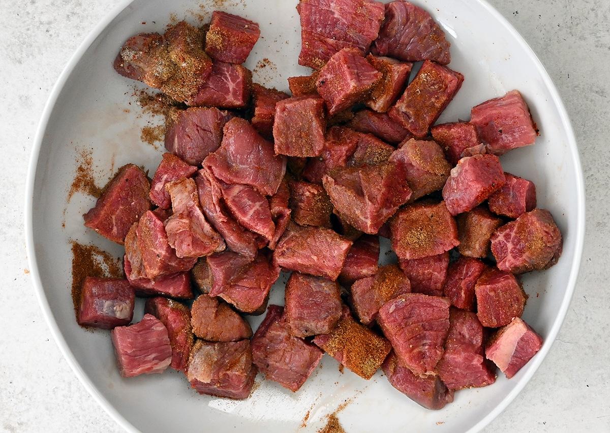 Chunks of raw beef sprinkled with seasoning are arranged in a white bowl on a light-colored surface, ready to become a hearty crock pot carne cuisada.