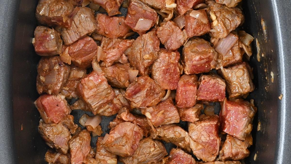 Cubed pieces of browned beef with chopped onions and seasonings simmering together in a pan for a classic crock pot carne cuisada.