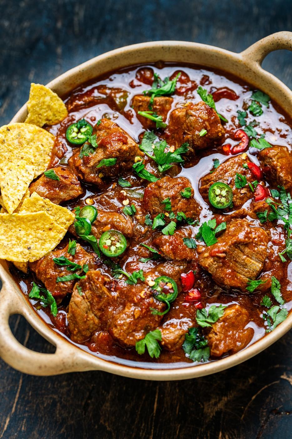 A bowl of spicy Crock Pot carne guisada beef stew garnished with sliced jalapeños and fresh cilantro, served with tortilla chips on the side. The rich, red sauce covers chunks of tender meat and vegetables.