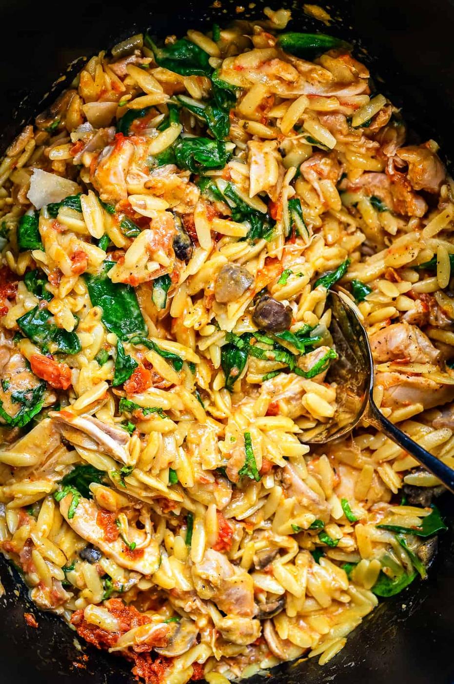 Close-up of creamy orzo pasta with spinach, tomatoes, mushrooms, and chicken, all blended together in a colorful and hearty meal. A spoon rests in the dish, ready to serve this comforting slow cooker favorite.