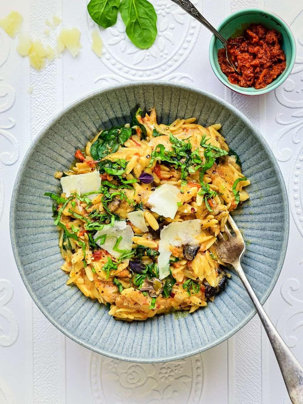 A bowl of creamy orzo pasta with spinach, mushrooms, shaved parmesan, and herbs is served with a fork. Nearby, sun-dried tomato spread and fresh spinach leaves add extra flavor to the white patterned surface.