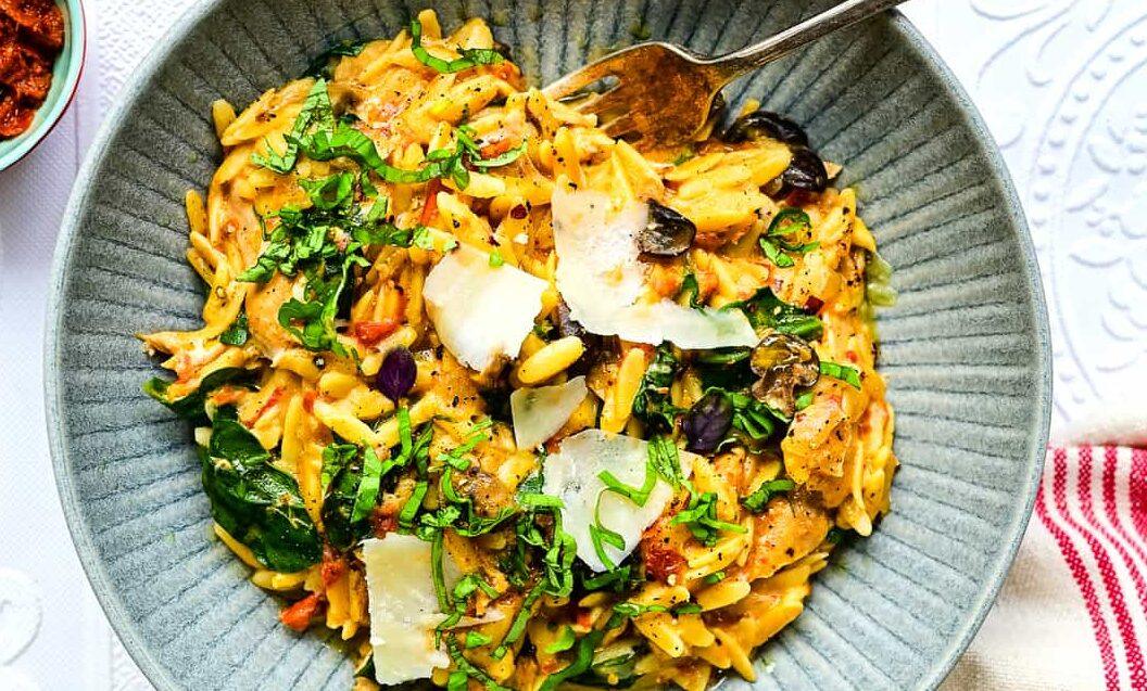 A gray bowl filled with creamy pasta, sun dried tomatoes, shaved parmesan, spinach, olives, and fresh herbs. A fork rests in the bowl, which sits on a red-striped napkin on a white textured surface.