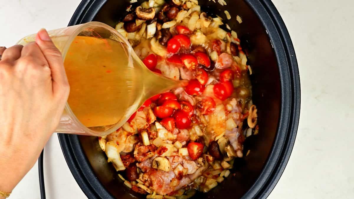 A hand pours broth from a measuring cup into a slow cooker filled with onions, tomatoes, mushrooms, chicken pieces, and sun dried tomatoes—perfect for making Crockpot Marry Me Chicken Orzo.