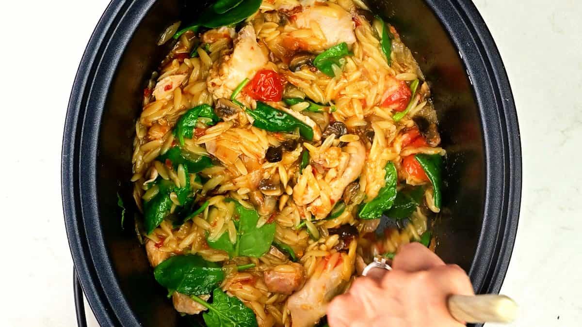A hand stirs a slow cooker brimming with creamy Crockpot Marry Me Chicken Orzo, mixed with spinach, mushrooms, and tomatoes in a savory sauce.