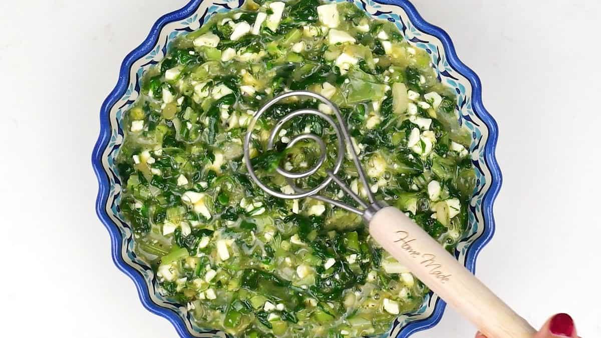 A blue-rimmed bowl filled with finely chopped green and white vegetables in liquid, perfect for a slimming Spinach Quiche, with a hand holding a pastry blender tool labelled Home Made above the mixture.