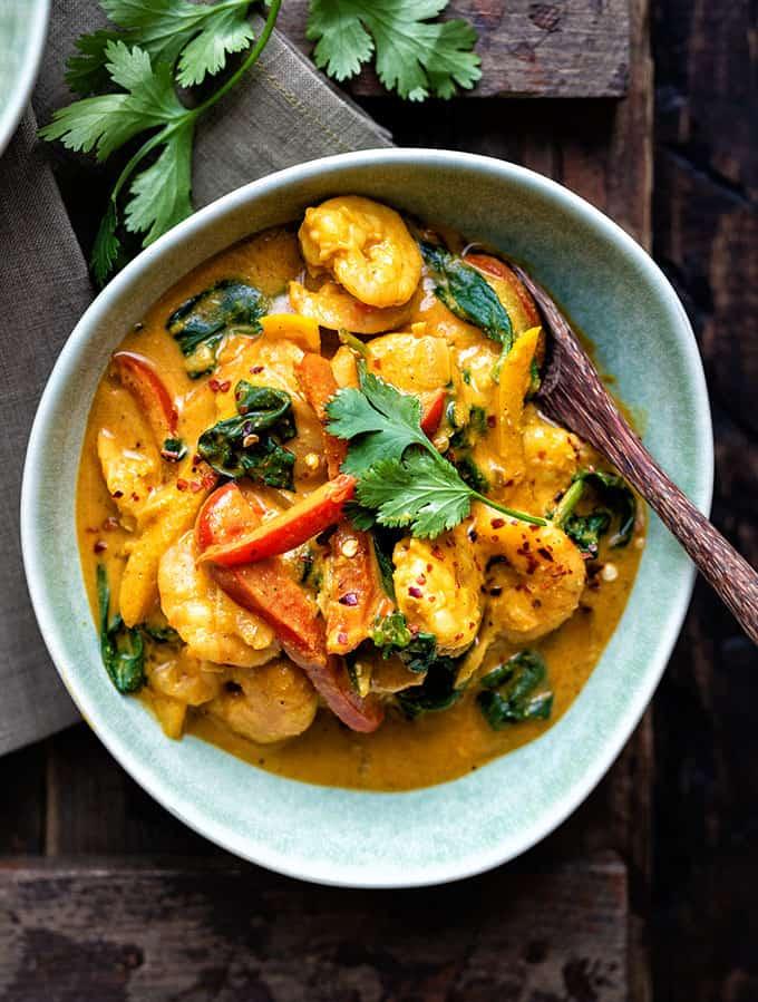 A bowl of prawn curry with spinach and red bell peppers in a yellow sauce, garnished with fresh cilantro, sits on a rustic wooden table beside a wooden spoon.