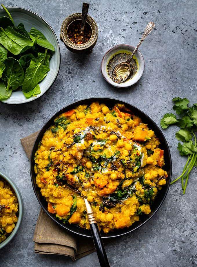 A bowl of hearty yellow curry with chickpeas, spinach, and carrots, garnished with seeds, sits on a gray surface. This vibrant vegan dish is complemented by fresh spinach, spices, cilantro, and a spoon arranged nearby.